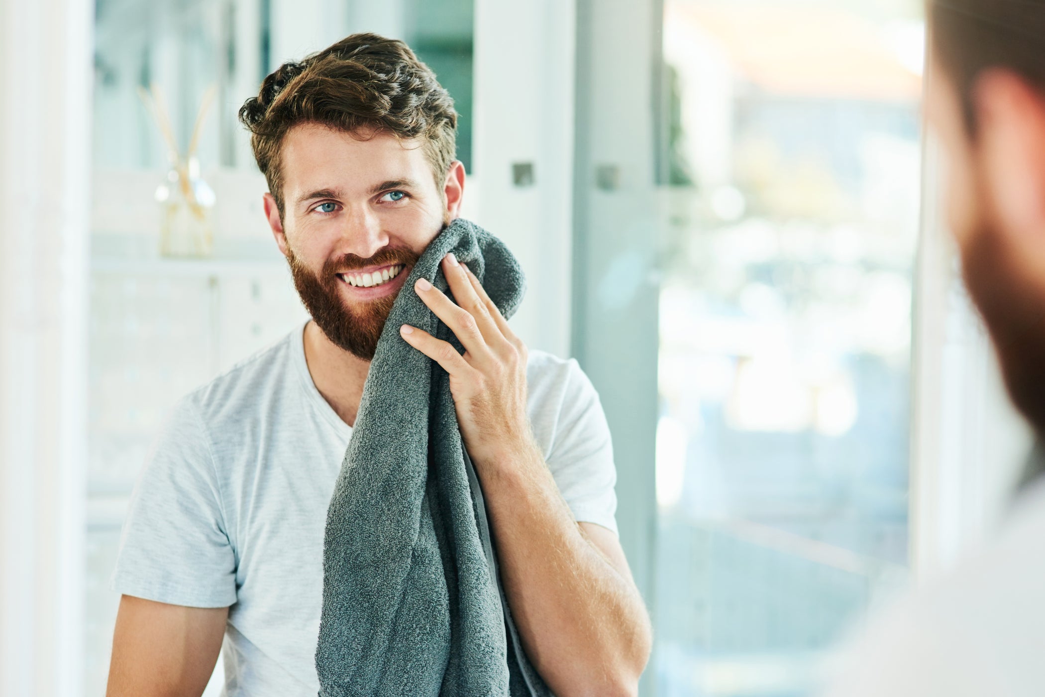 Giovane uomo con capelli castani e barba si asciuga il viso con un asciugamano in bagno