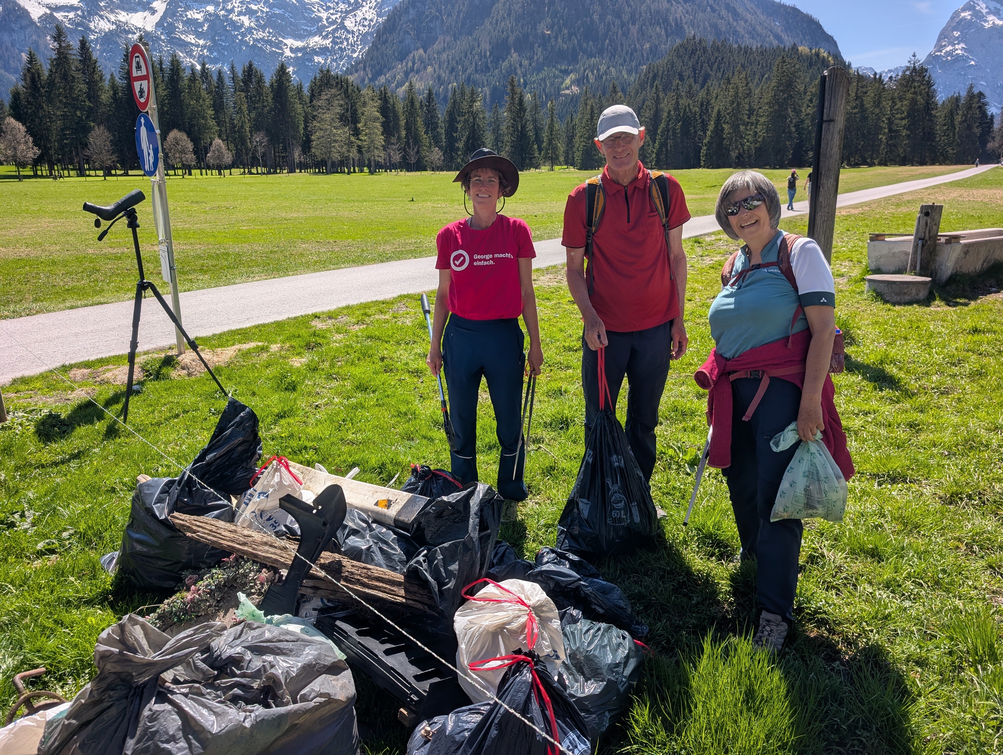 Drei Personen stehen auf einer Wiese vor Berglandschaft mit Tüten vor sich 
