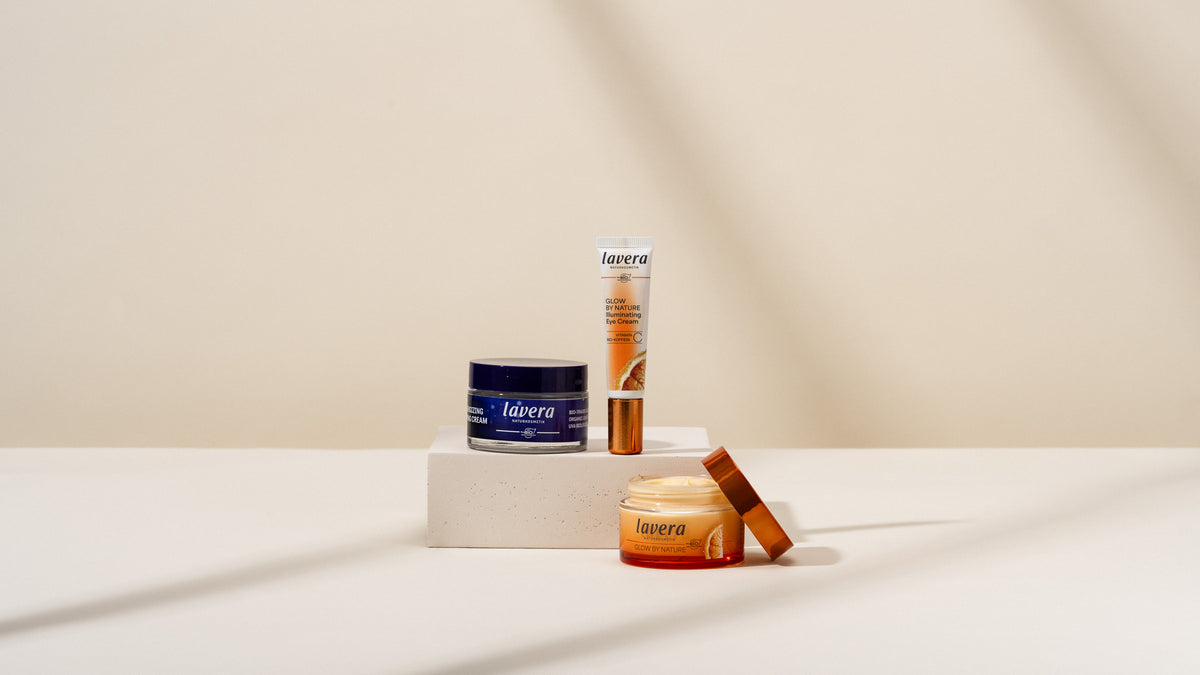 lavera Glow by Nature Produkte auf einem beigen Podest: links ein dunkelblauer Tiegel, in der Mitte eine Augencreme-Tube und rechts ein geöffneter orangefarbener Tiegel mit Zitrusmotiv.