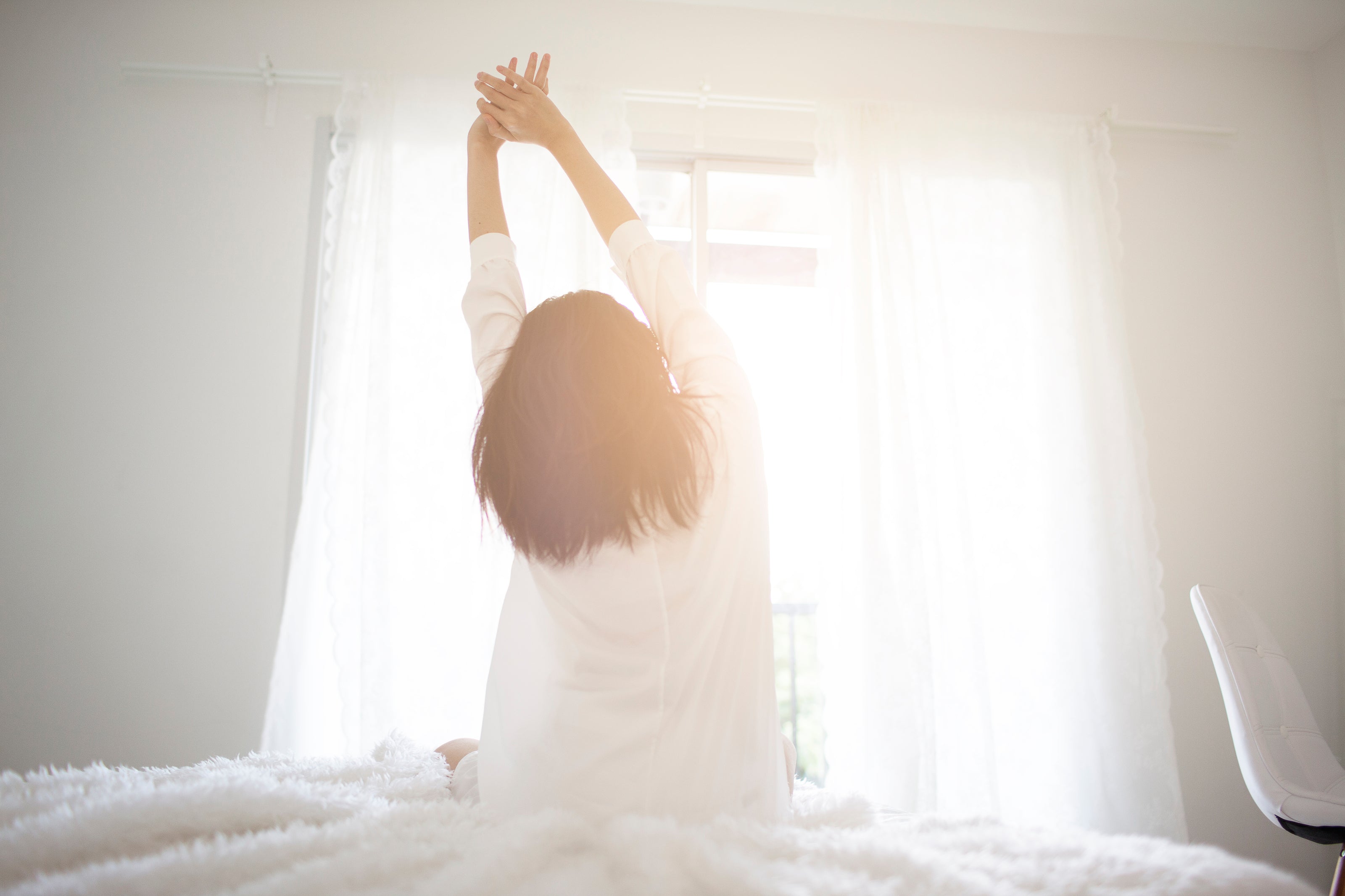 Donna seduta di spalle alla camera su un letto e si allunga – la luce del sole entra dalla finestra