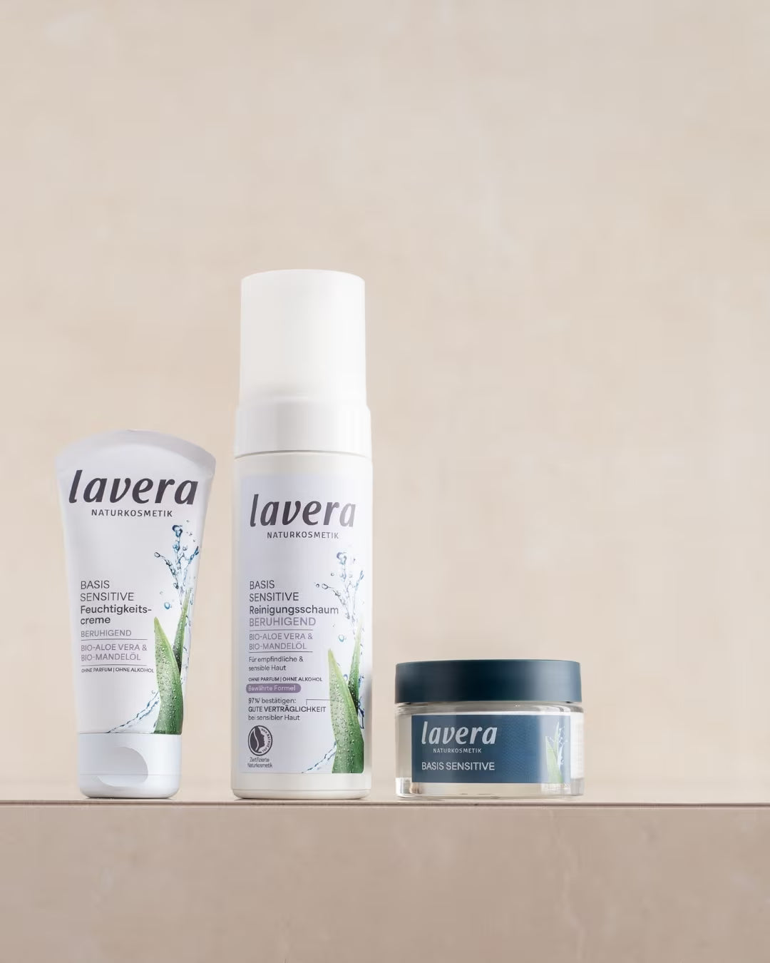 Drei lavera Basis Sensitive Pflegeprodukte – Feuchtigkeitscreme, Reinigungsschaum und Gesichtscreme – stehen nebeneinander auf einem beigefarbenen Podest.