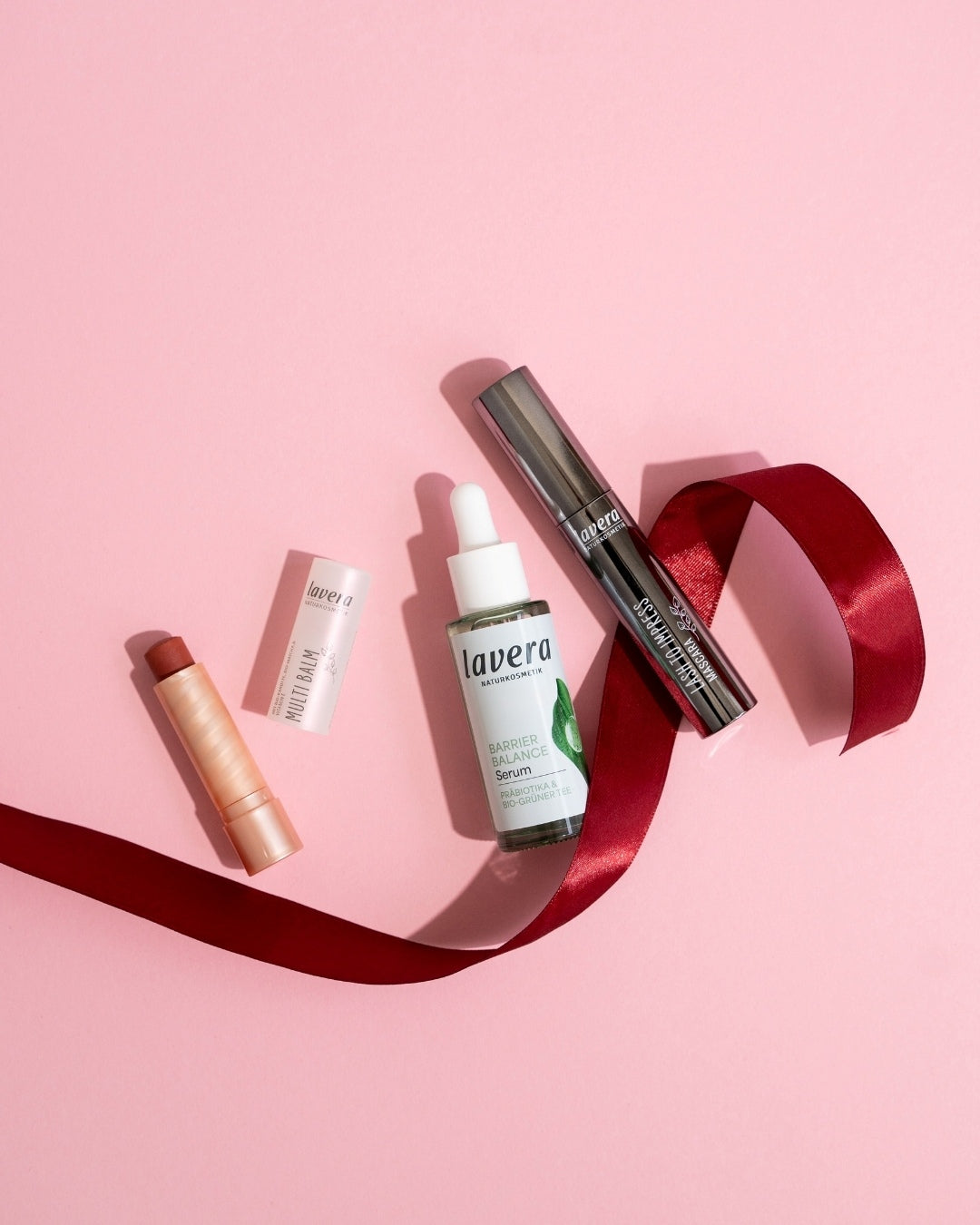 Multi Balm, Barrier Balance Serum und Lash to Impress Mascara auf einem roten Geschenkband