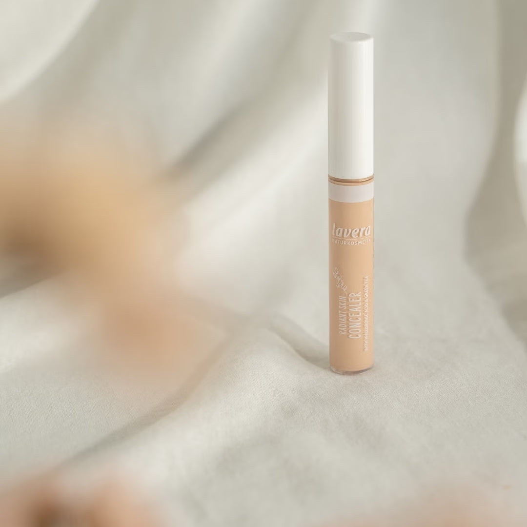 Un correttore liquido per la pelle lavera è appoggiato su un tessuto chiaro davanti a uno sfondo drappeggiato delicatamente, con accenti beige sfocati.