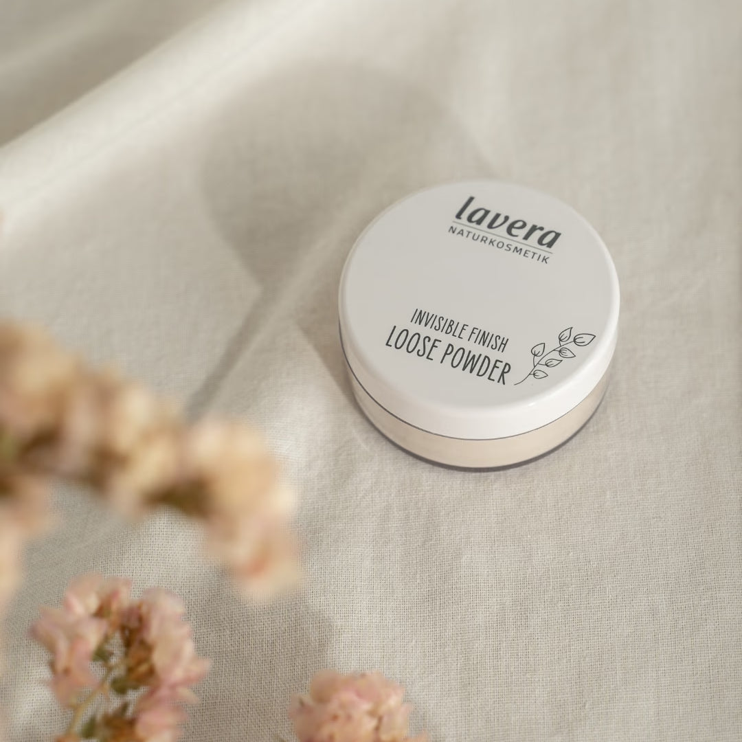 lavera Invisible Finish Loose Powder liegt auf hellem Leinenstoff, umgeben von unscharfen rosafarbenen Blüten.