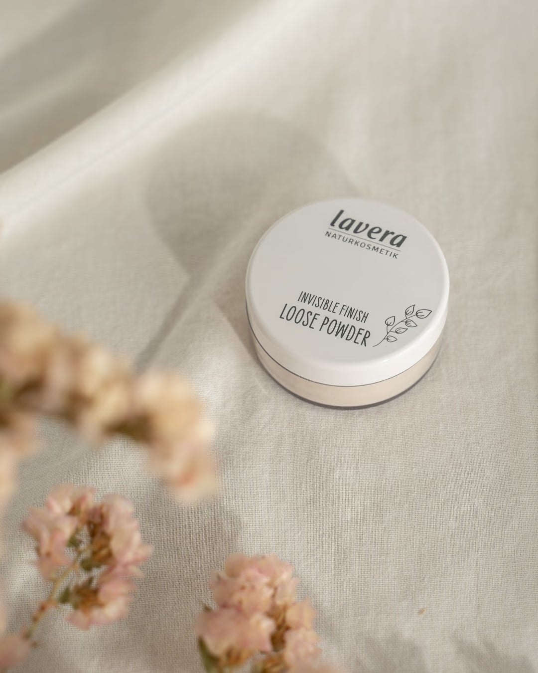 lavera Invisible Finish Loose Powder liegt auf hellem Leinenstoff, umgeben von unscharfen rosafarbenen Blüten.
