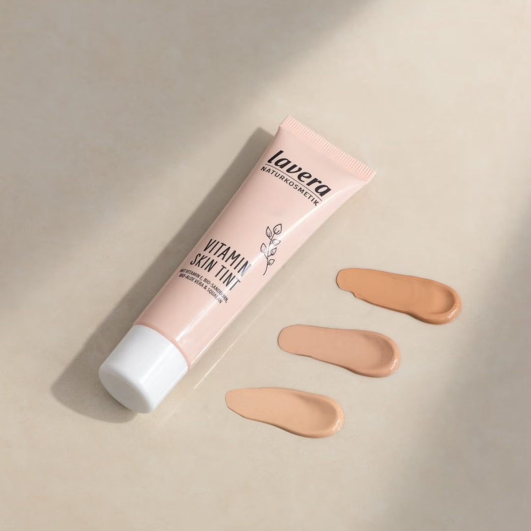 Eine Tube lavera Vitamin Skin Tint liegt neben drei unterschiedlich hellen Farbtupfern auf einer beigefarbenen Oberfläche.