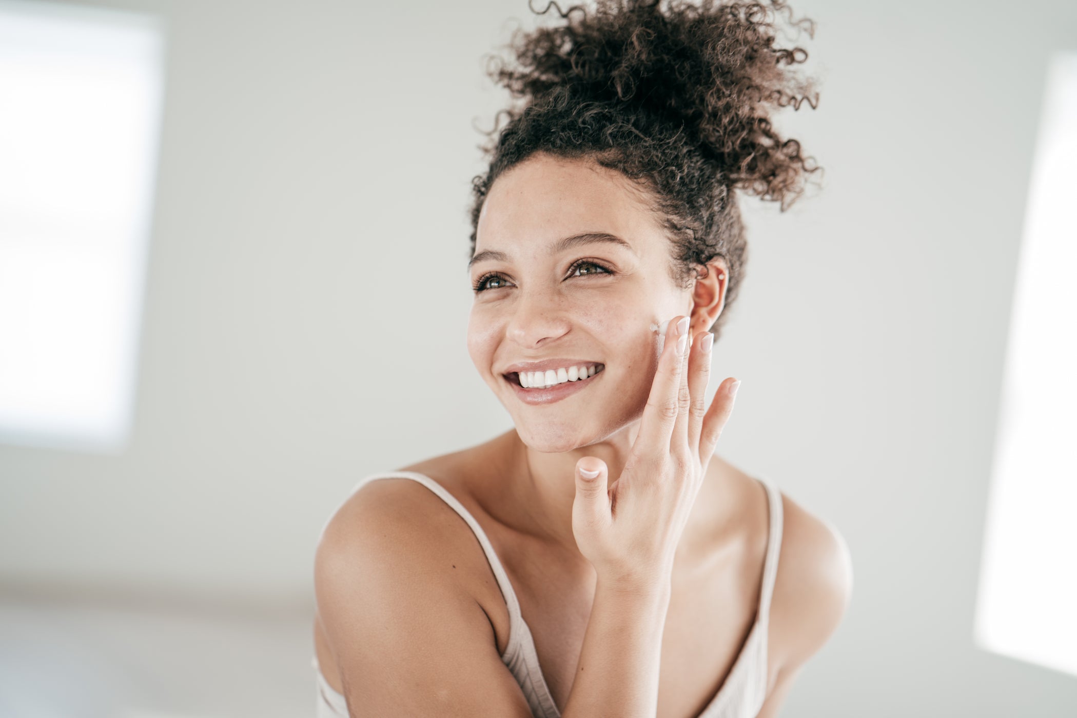 Giovane donna con capelli scuri e ricci sorride e applica crema sulle guance con il dito