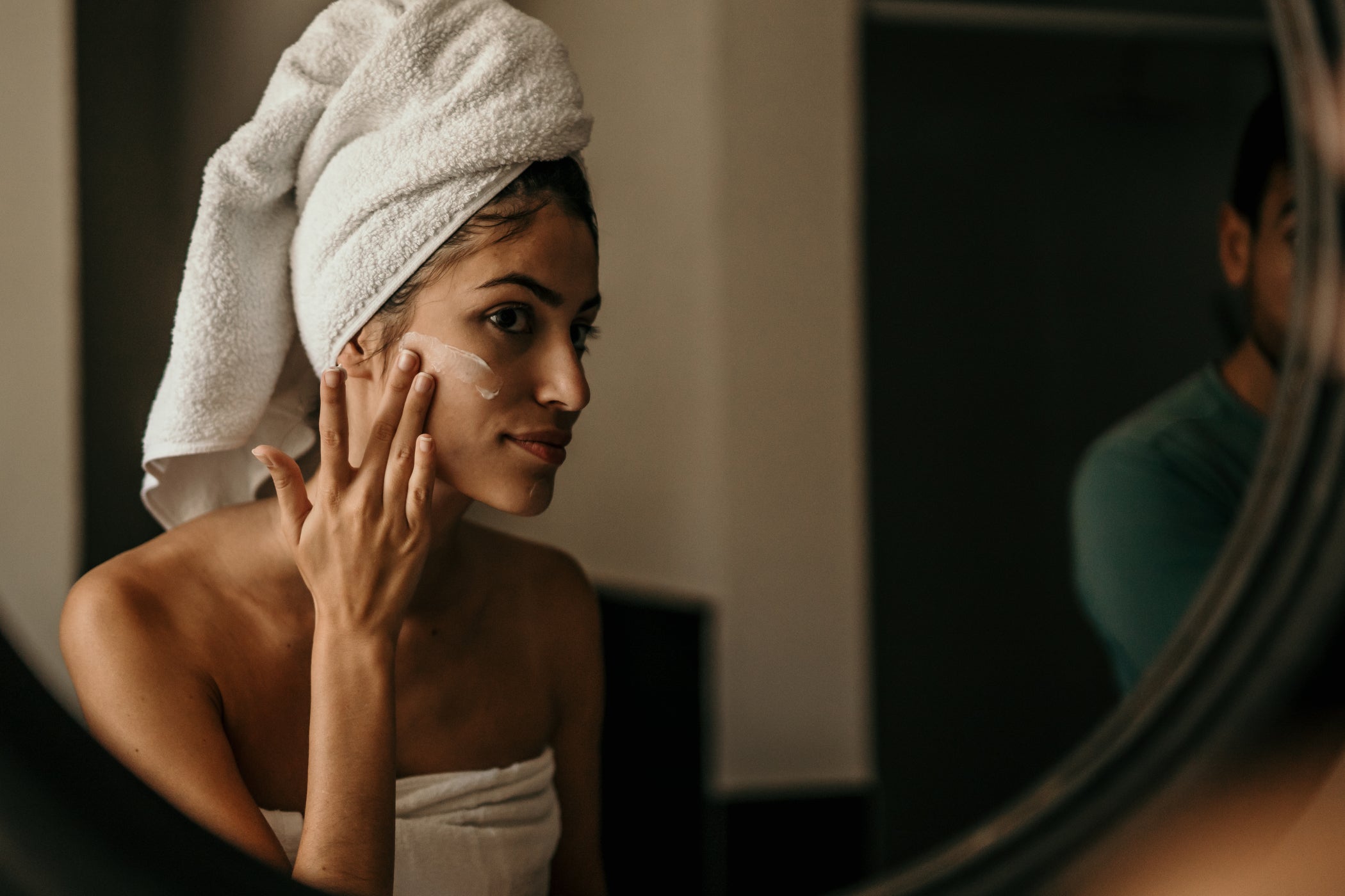 Donna con turbante asciugamano applica crema viso, specchio in bagno