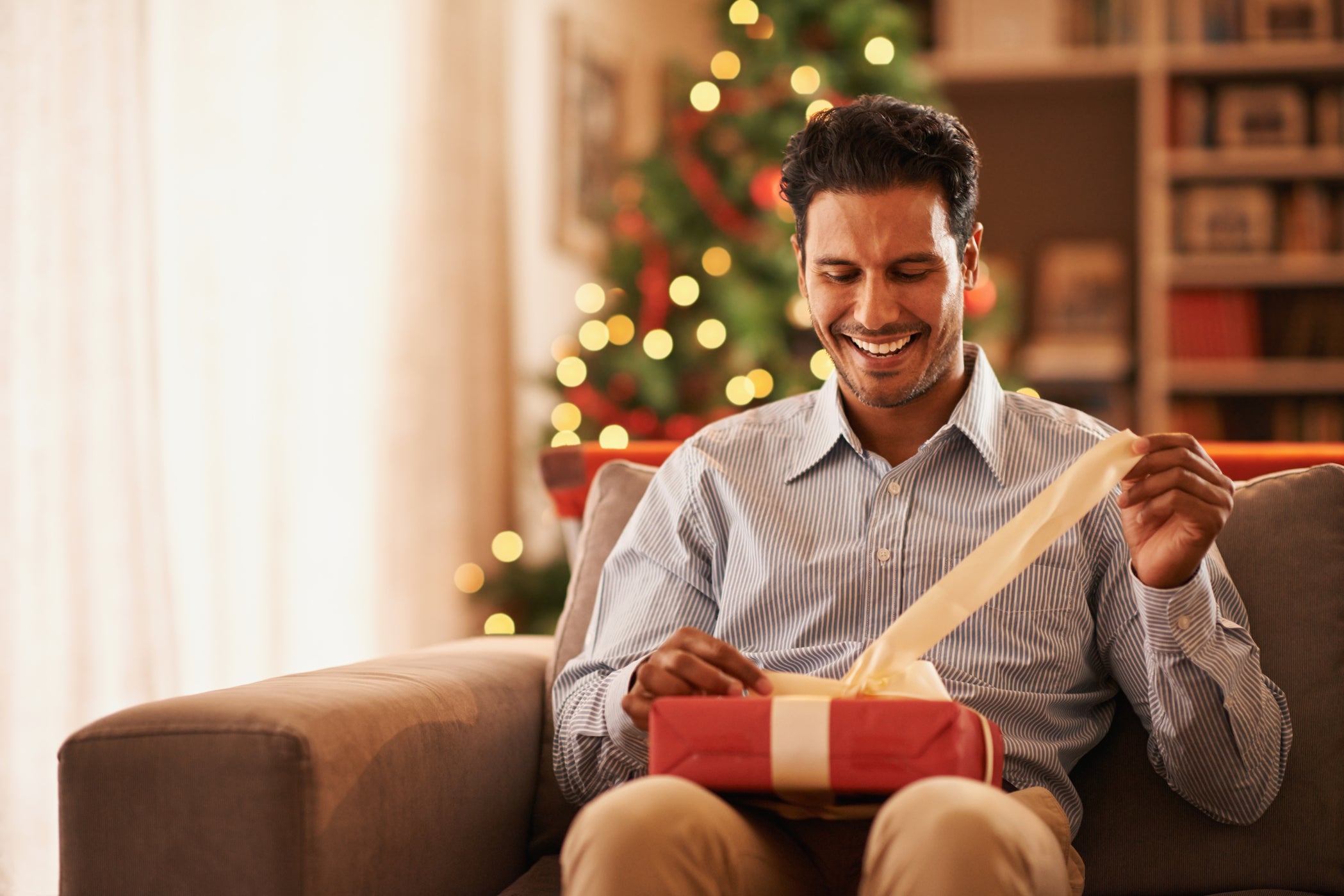 Uomo seduto su un divano scarta un regalo con albero di Natale sullo sfondo