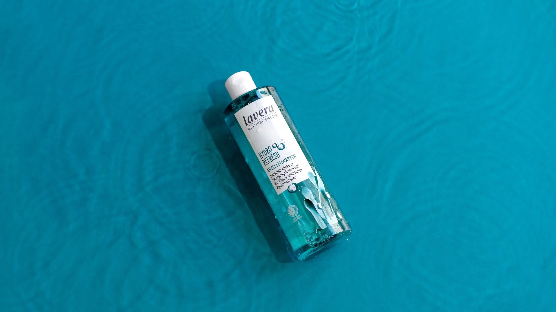 Un flacone di acqua micellare lavera Hydro Refresh è appoggiato su una superficie strutturata di un turchese brillante, alla luce del sole.