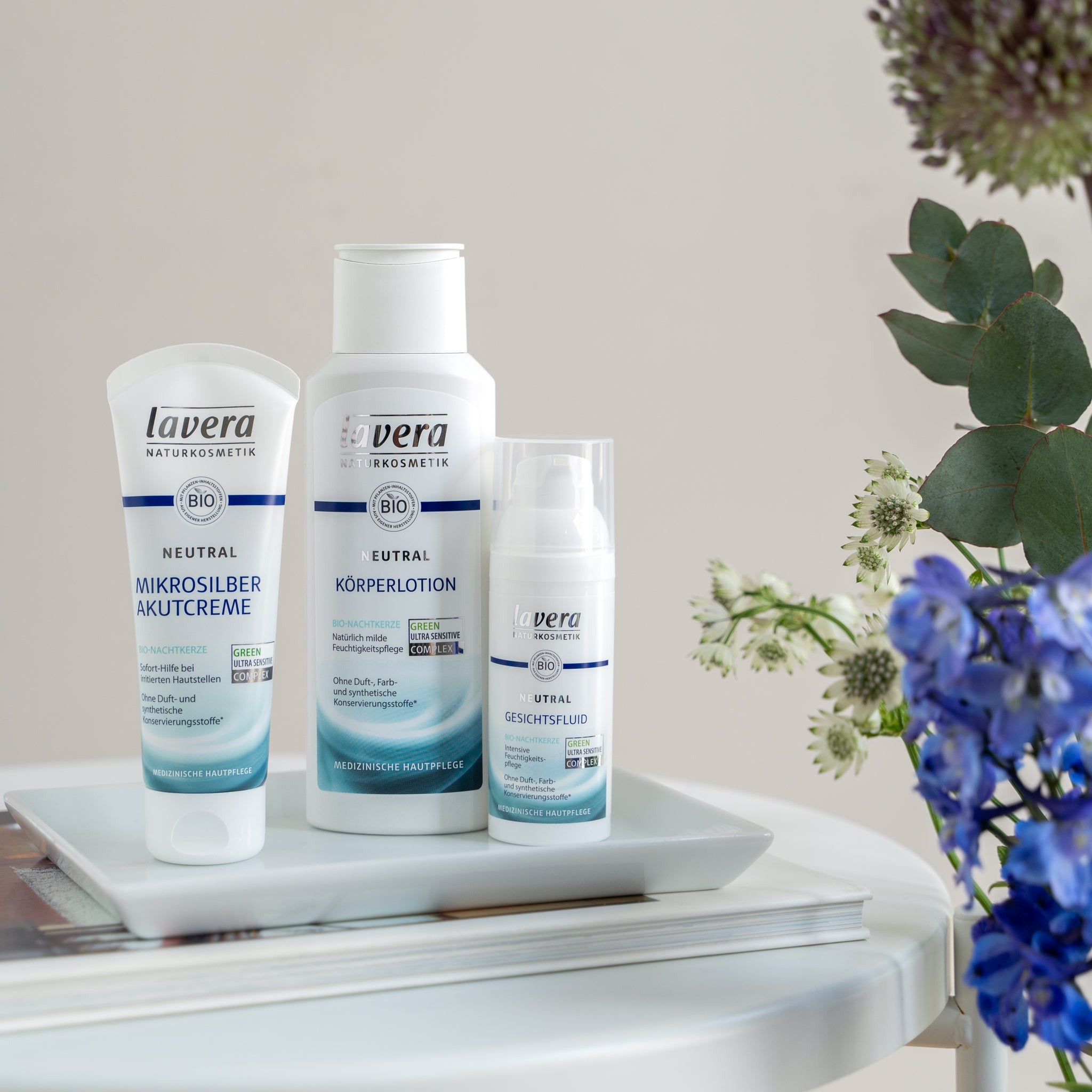 lavera Neutral prodotti di trattamento dermatologico con microargento: crema d'emergenza, lozione corpo e fluido viso, disposti decorativamente con fiori su tavolo bianco
