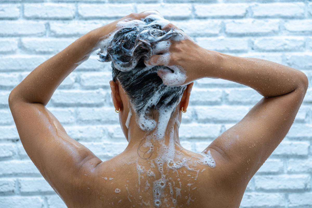 Persona femminile di spalle alla camera si lava i capelli con shampoo molto schiumoso