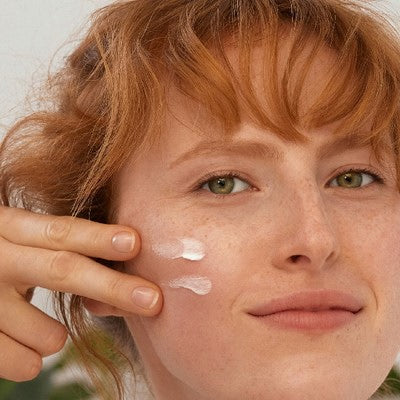 Donna dai capelli rossi che applica crema sulla guancia, due punti visibili