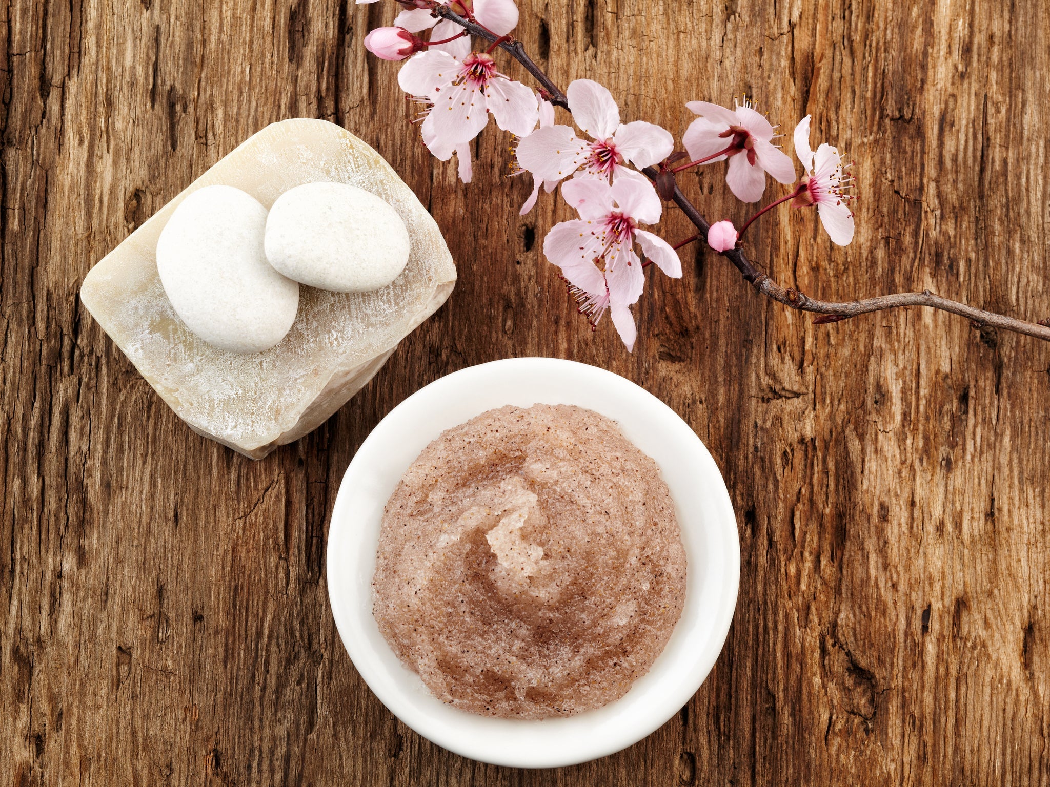 Schale mit angerührtem Zuckerpeeling, daneben Dekoration mit Steinen und Kirschblütenzweig auf Holzuntergrund.