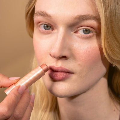 Giovane donna con capelli biondi che sorride alla telecamera applicando Multi Balm sulle labbra