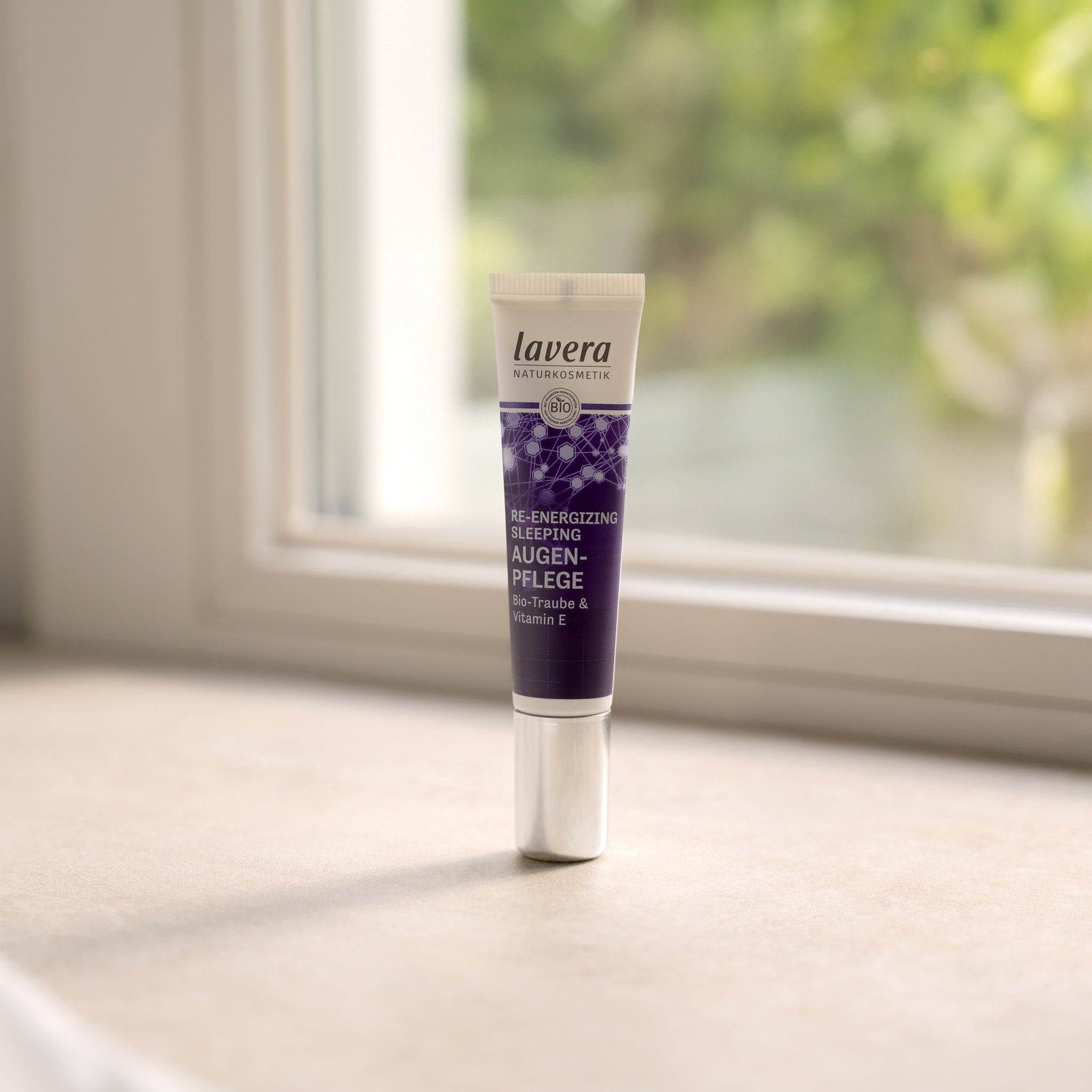 Un piccolo tubetto viola con chiusura argentata del prodotto per il contorno occhi lavera Naturkosmetik Re-Energizing Sleeping posizionato su un davanzale alla luce del giorno, verde sfocato sullo sfondo