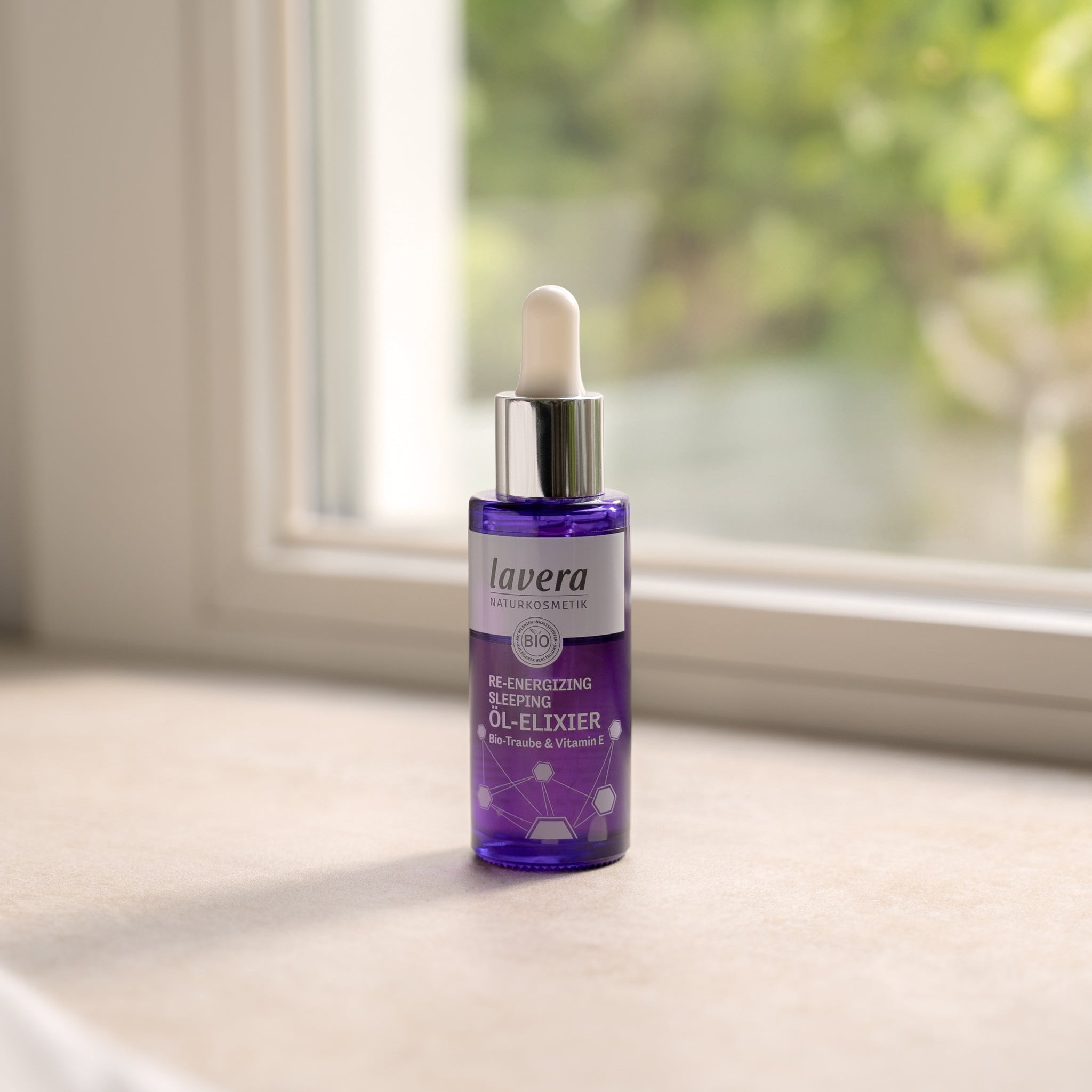 Una bottiglia di vetro viola con pipetta dell'olio elisir lavera Naturkosmetik Re-Energizing Sleeping in piedi su un davanzale alla luce del giorno soffusa, con verde sfocato sullo sfondo