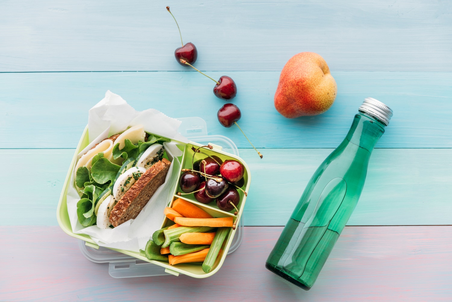 lunch-box-nachhaltig-stock-magazin-rezepte Porta pranzo con un panino e verdure crude colorate accanto a una bottiglia di vetro verde e frutta fresca su un tavolo di legno pastello
