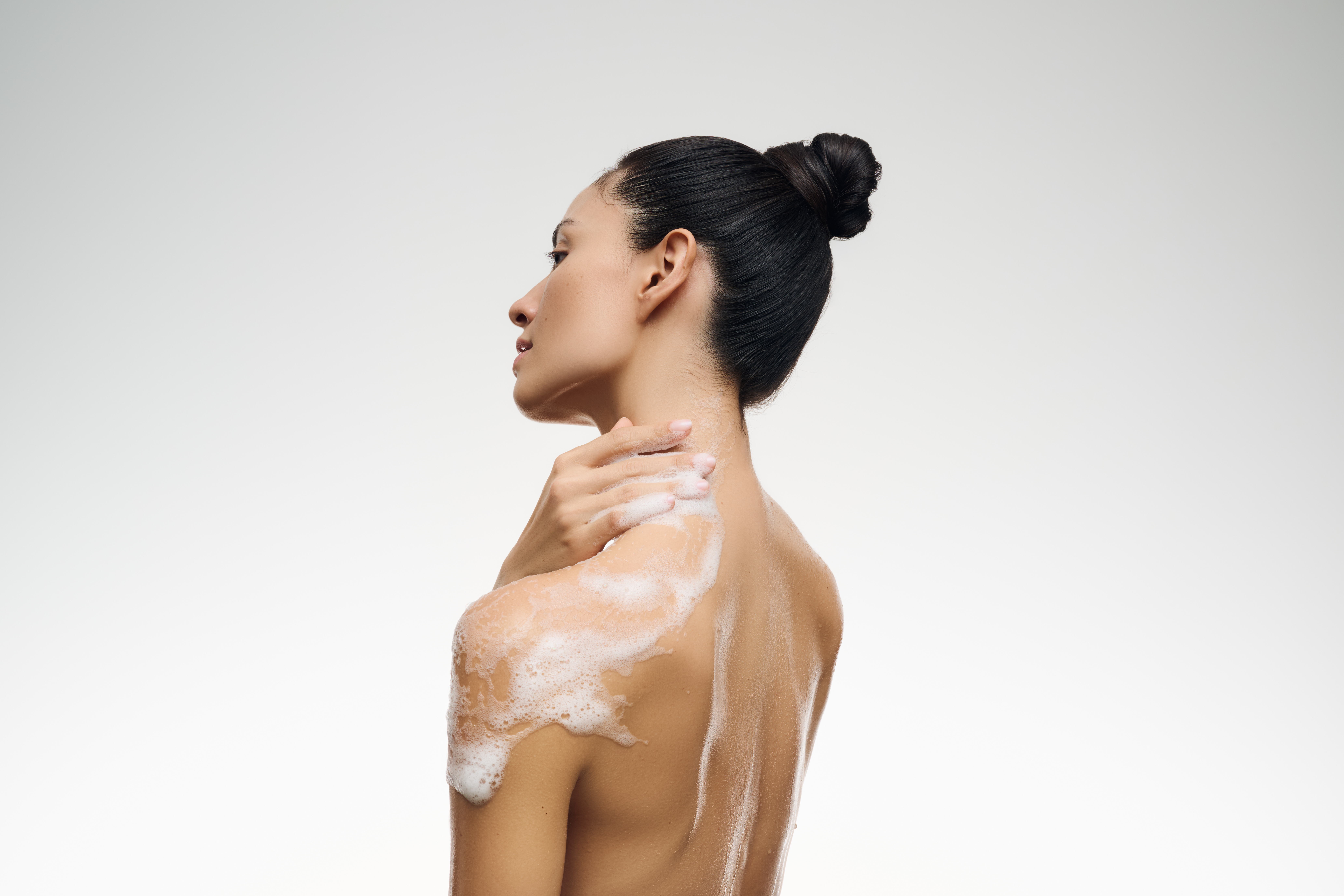 Frau mit dunklen Haaren und Dutt macht sich schäumendes Duschgel auf die nackte Haut